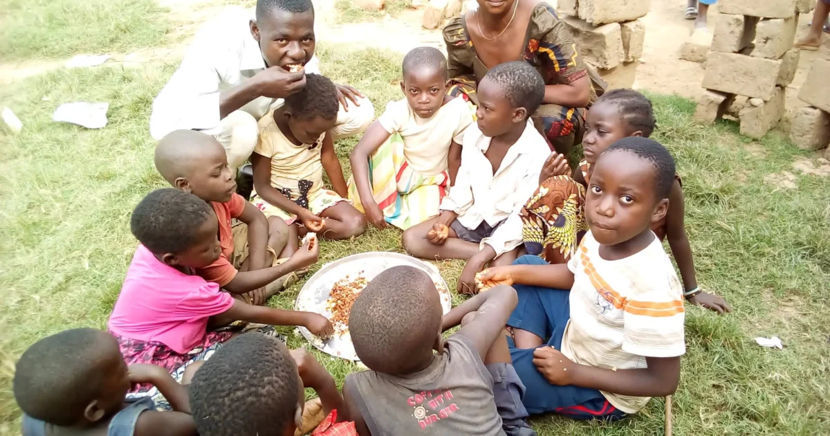 Groupe d'enfants et d'adultes assis en cercle sur l'herbe partageant un repas sur un plateau métallique lors d'une activité communautaire.