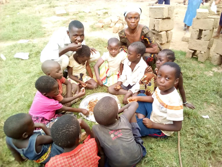 Groupe d'enfants et d'adultes assis en cercle sur l'herbe partageant un repas sur un plateau métallique lors d'une activité communautaire.